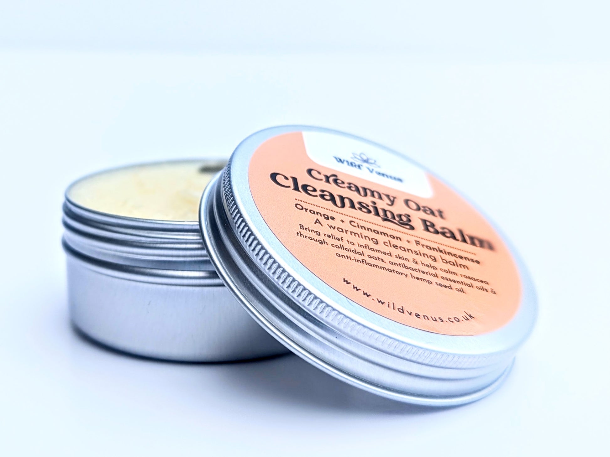 An open tin of Creamy Oat Cleansing Balm with the tin resting on the open pot. The tin lid is in the foreground and thhe product is set against a white background. 