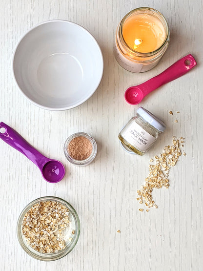 A photo from above shows some oats, a lit candle, a pot of clay face mask on its side, and another with the top off. There are 2 measuring spoons and a bowl. 