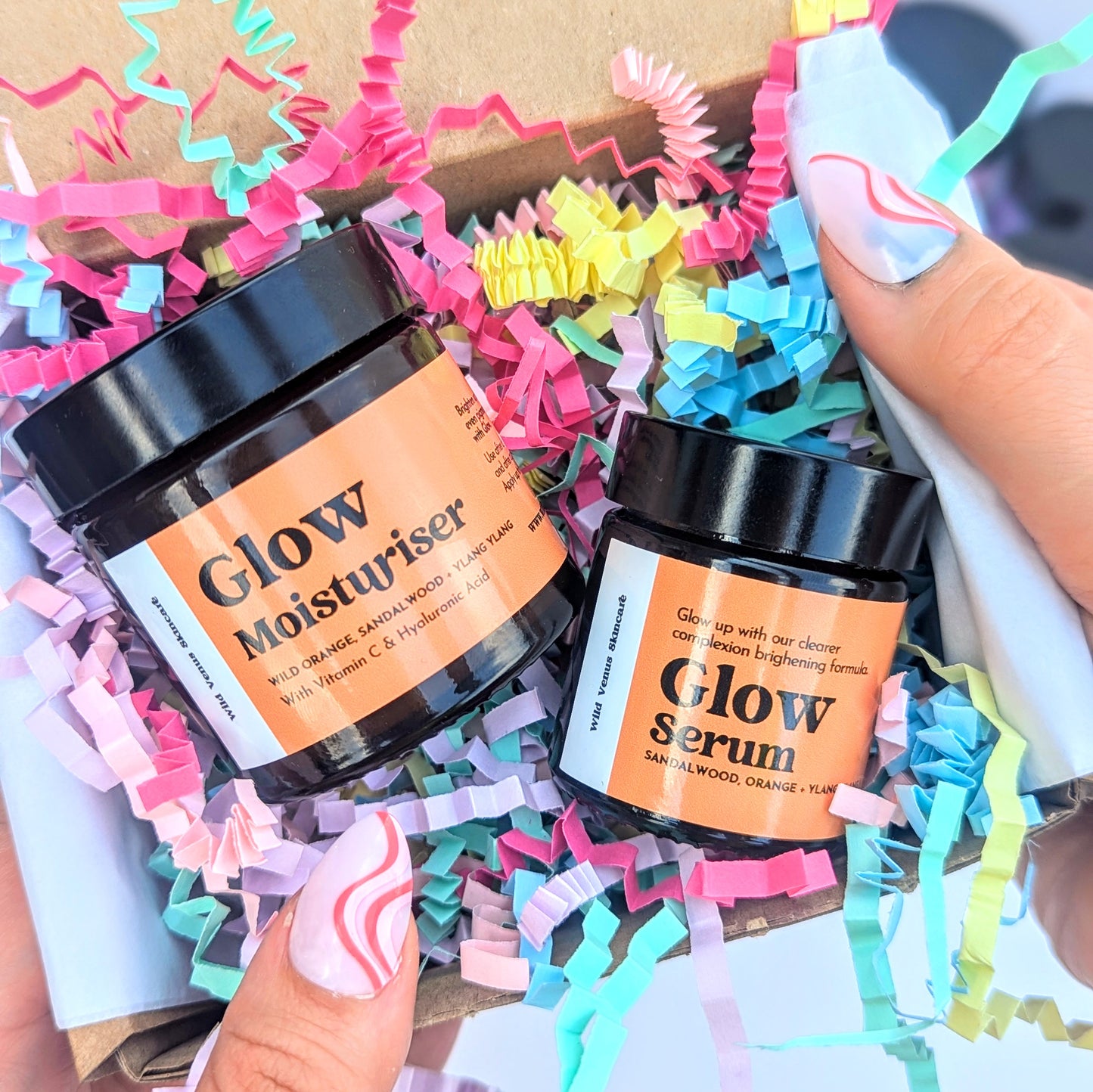 Ladies hands are shown holding a gift box with colourful crinkle paper in and skin care products. The skincare products are the Glow serum and moisturiser. 