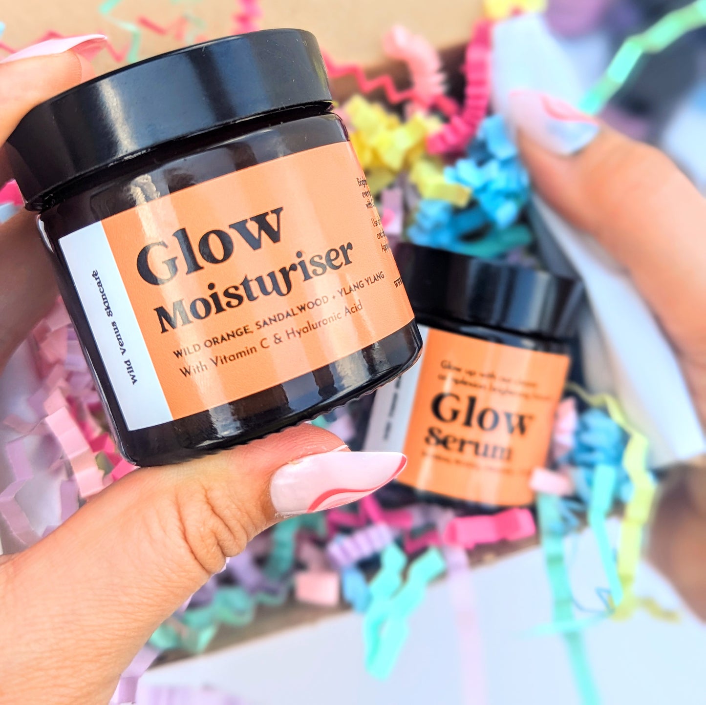 A hand is shown holding a Glow Moisturiser up to the camera. The other hand shows a gift box, full of brightly coloured shredded paper and the Glow Serum is presented inside. 