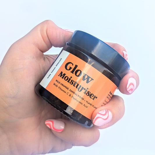 A ladies hand is shown graping an amber pot of Glow Moisturiser. The pot is tilted and there is a shine on the surface of the bright orange label. 