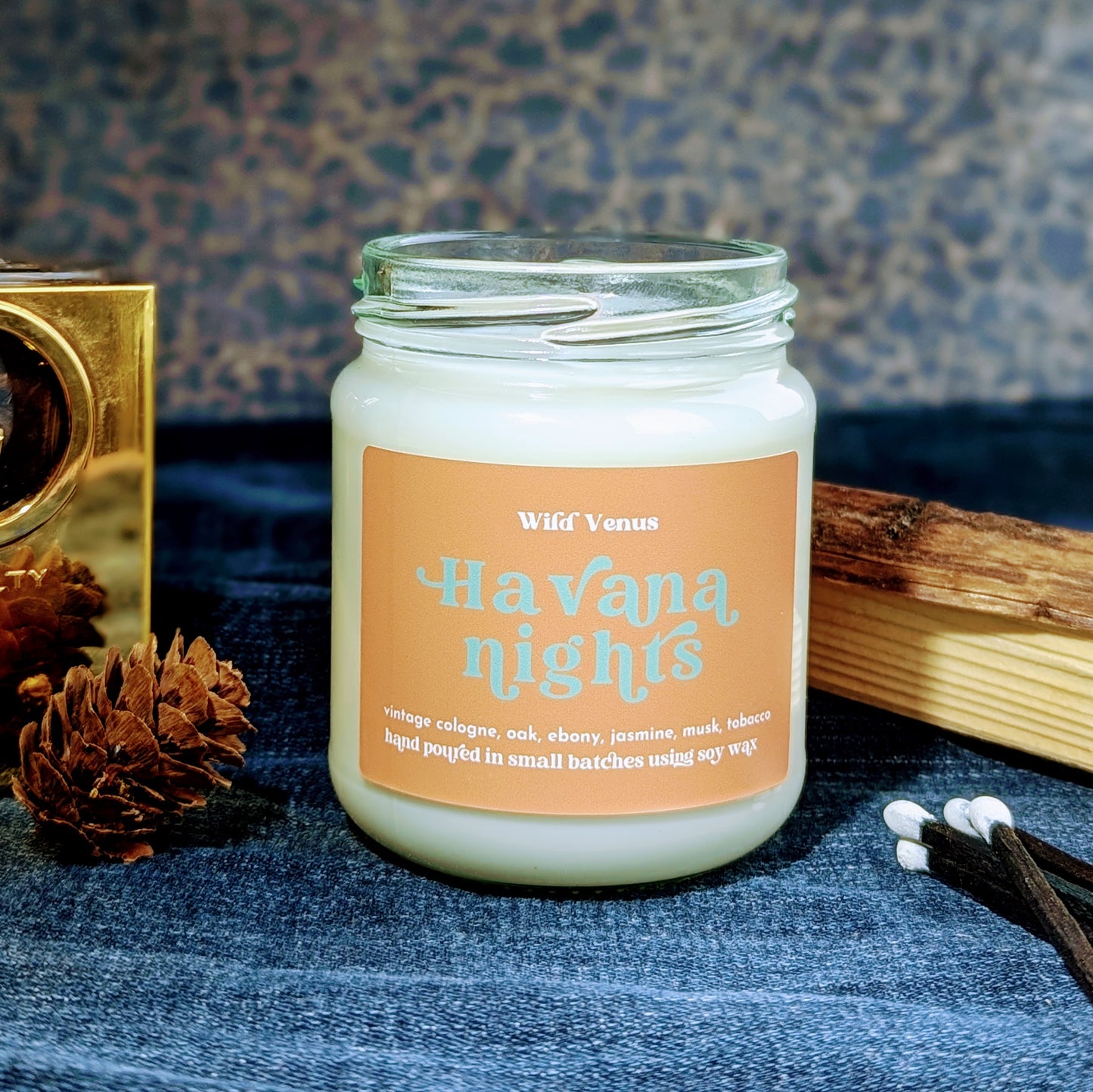 A soy candle called Havana Nights is on some dark blue denin. Beside it is some wood, a pinecone and some matches.