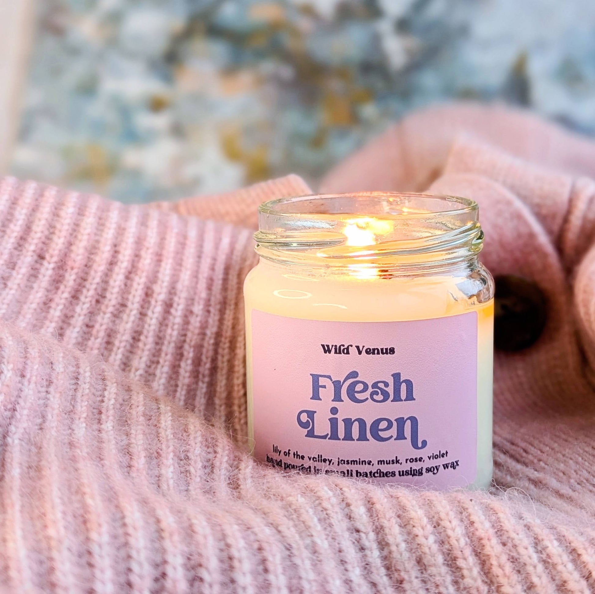 a lit fresh linen scented candle is shown nestled in a cosy pink sweater. 