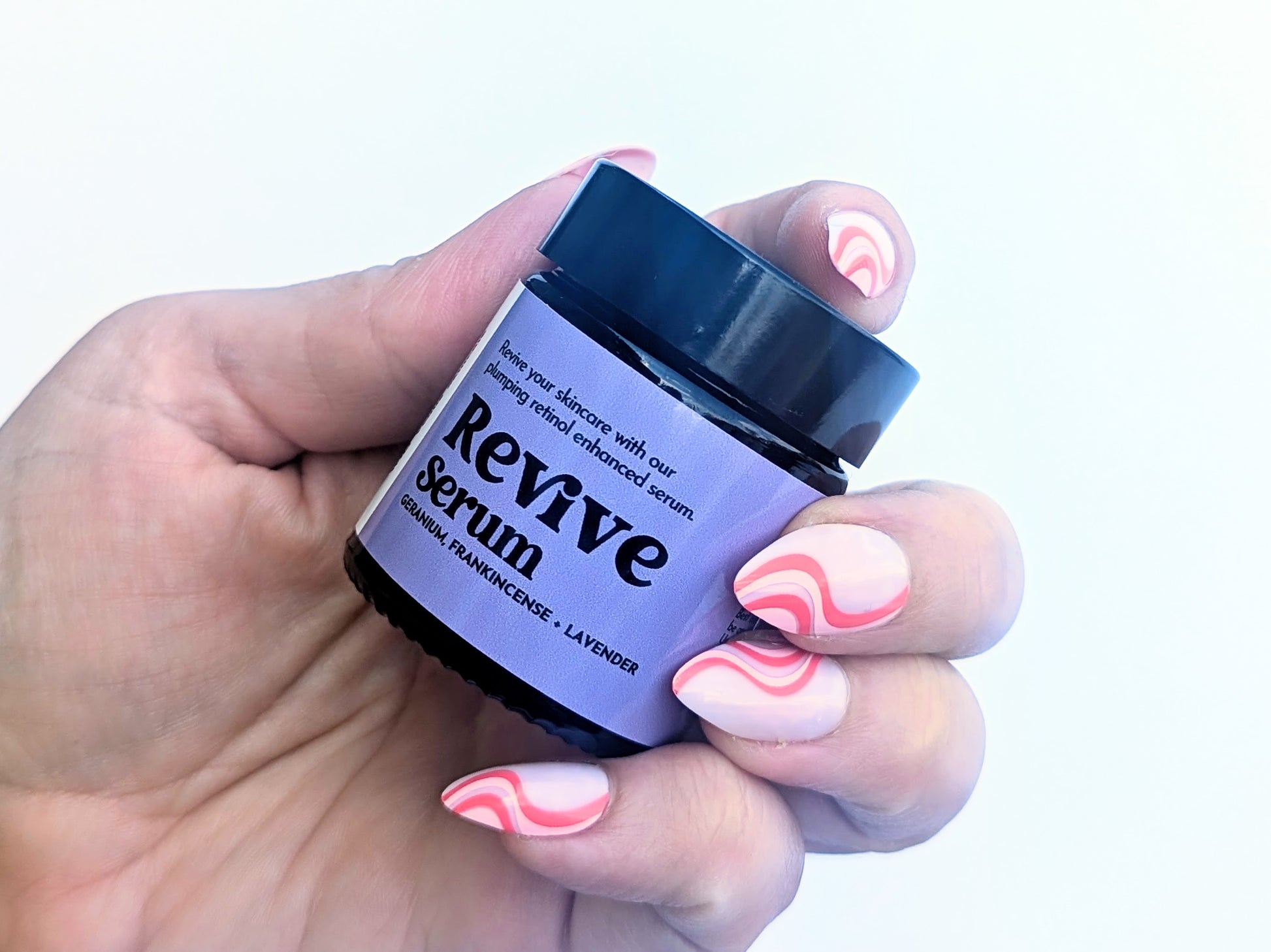 A manicured hand holds a small pot of the Revive serum against a white background. 