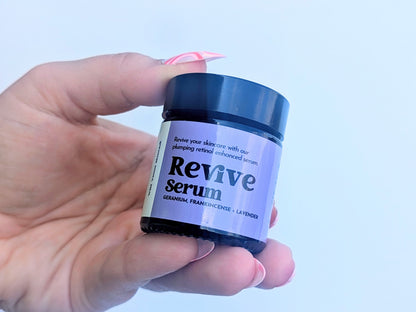 A manicured hand casually holds a small pot of the Revive serum against a white background.