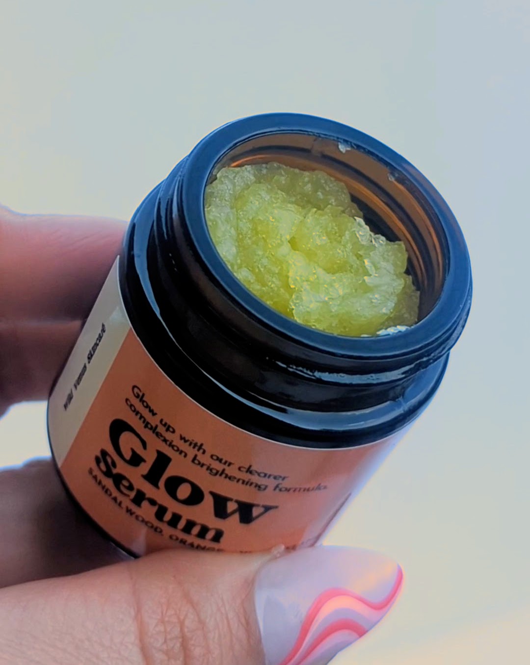 A hand tilts an open pot of Glow serum towards the viewer, revealing a golden solid oil inside. 