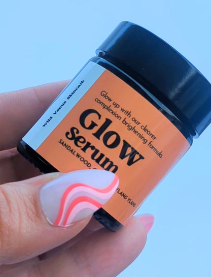 A manicured hand delicately holds the small pot of Glow Serum, the thumbnail slightly obscures the lettering. The serum is held up against a light blue background. 