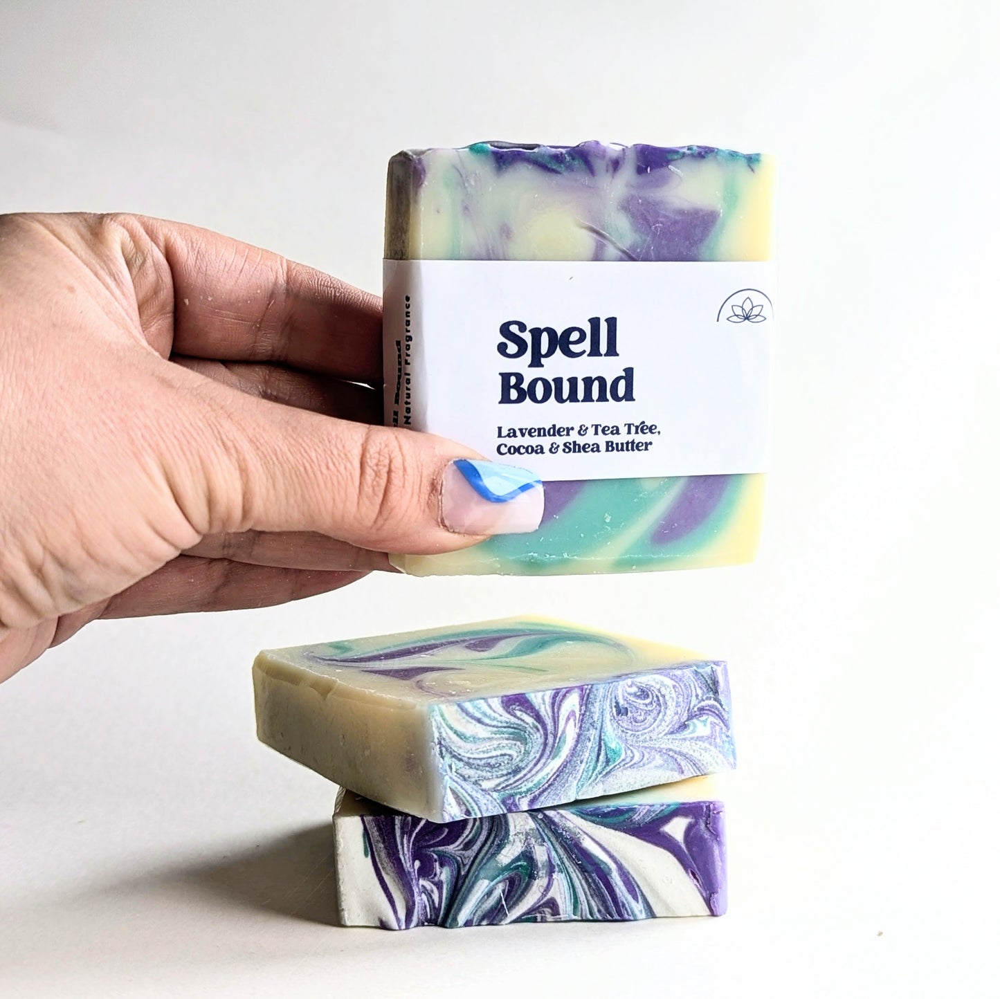 A hand picks up a bar of Spell bound lavender and tea tree soap from a pile of 3 soap bars.