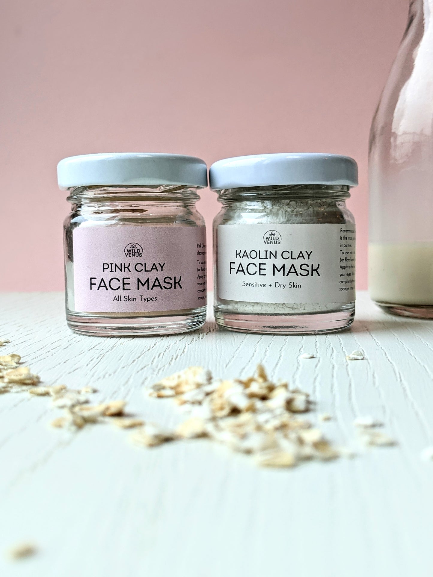 A pink clay and kaolin clay on a white table with a few oats scattered in the foreground. There is some oat milk in a small bottle by the side. 