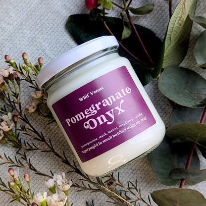 Square photo of a pomegranate soy candle on its side surrounded by foliage and some dainty flowers.