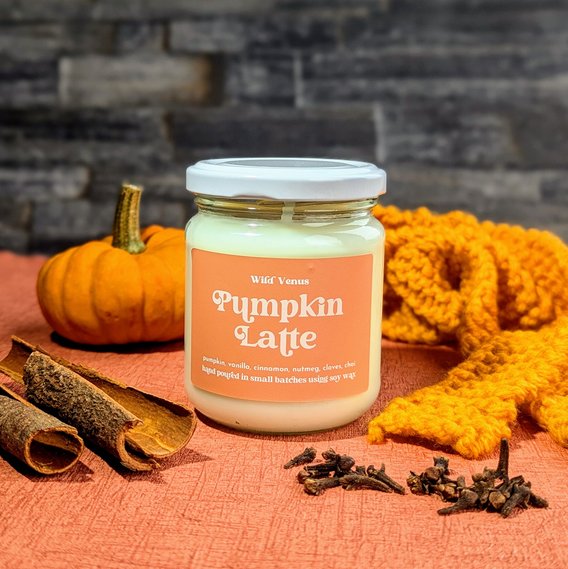 A candle with the Pumpkin Latte label on, it it on an orange surface with a slate brick wall background. In the foreground is cinnamon sticks, cloves, a mini pumpkin and an orange scarf.