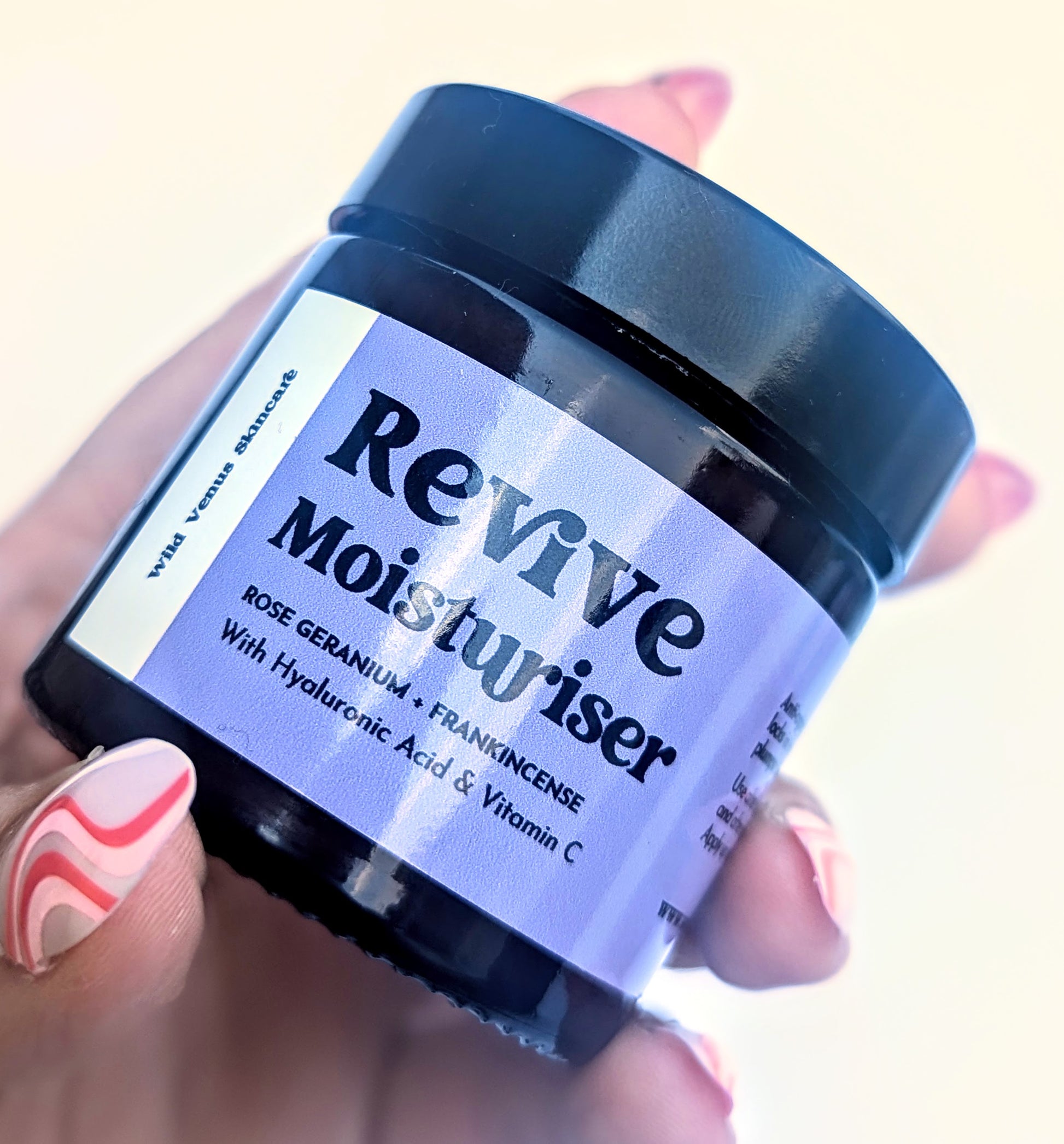 A manicured hand holds a pot of the Revive Moisturiser. 