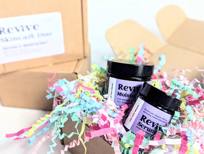 A Revive Skincare Duo Gift Box is presented close to the the viewer, the products are sitting up in the box, held in place by mounds of bright and fun shredded paper. Behind this gift box and out of focus are two more gift boxes stacked on top of each other. 