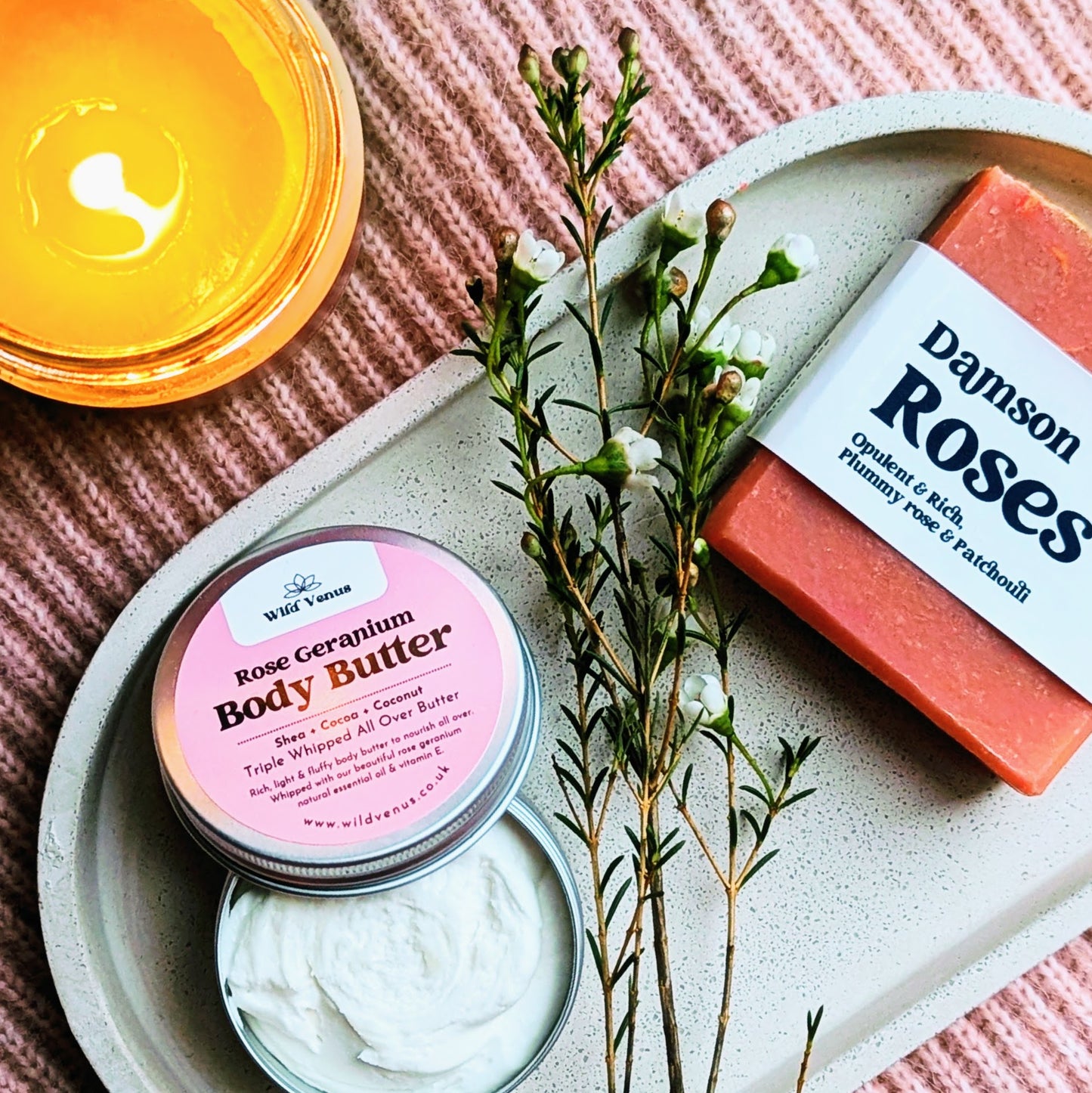 A photograph shows a lit candle, soap and a body butter from above. There's a small sprig of flowers.