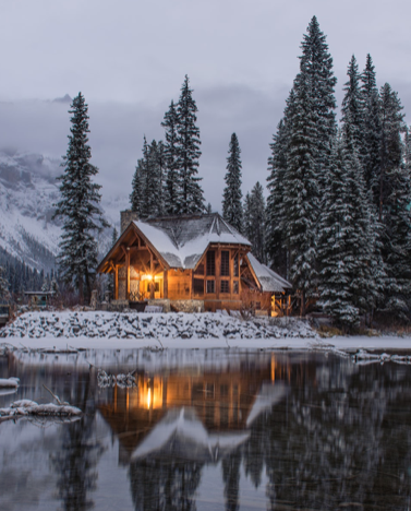 A beautiful lake side, log cabin at dusk surrounded by snow tipped trees and hills. 