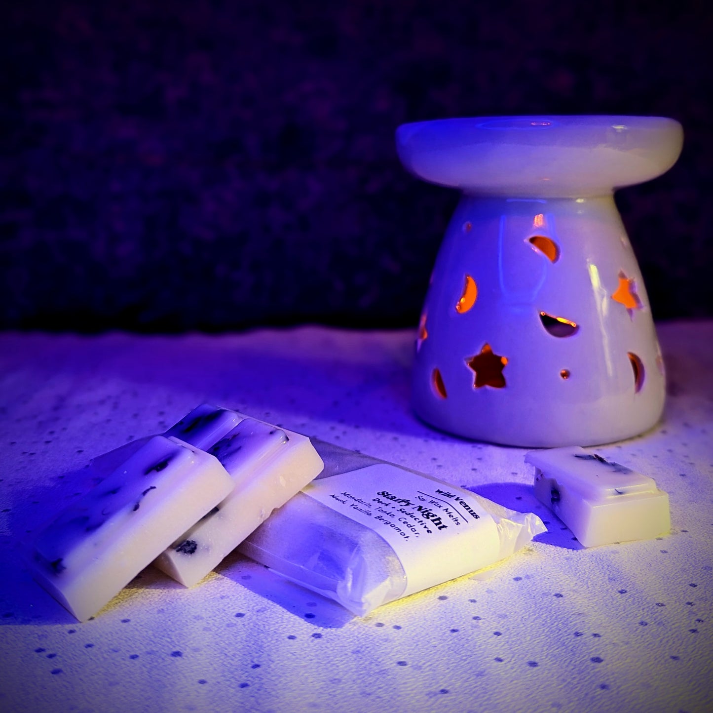 A starry night botanical soy wax melt has been snapped and is left in chunks on top of another wrapped wax melt bar. A white wax warmer is in the background with a warm glow.