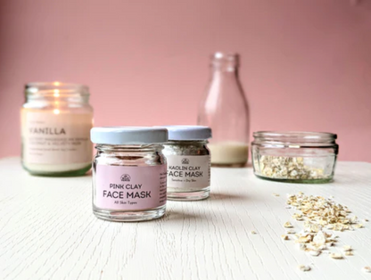 A table is set up with a pink clay, kaolin clay, oat milk and oats, A vanilla candle is lit in the background. 