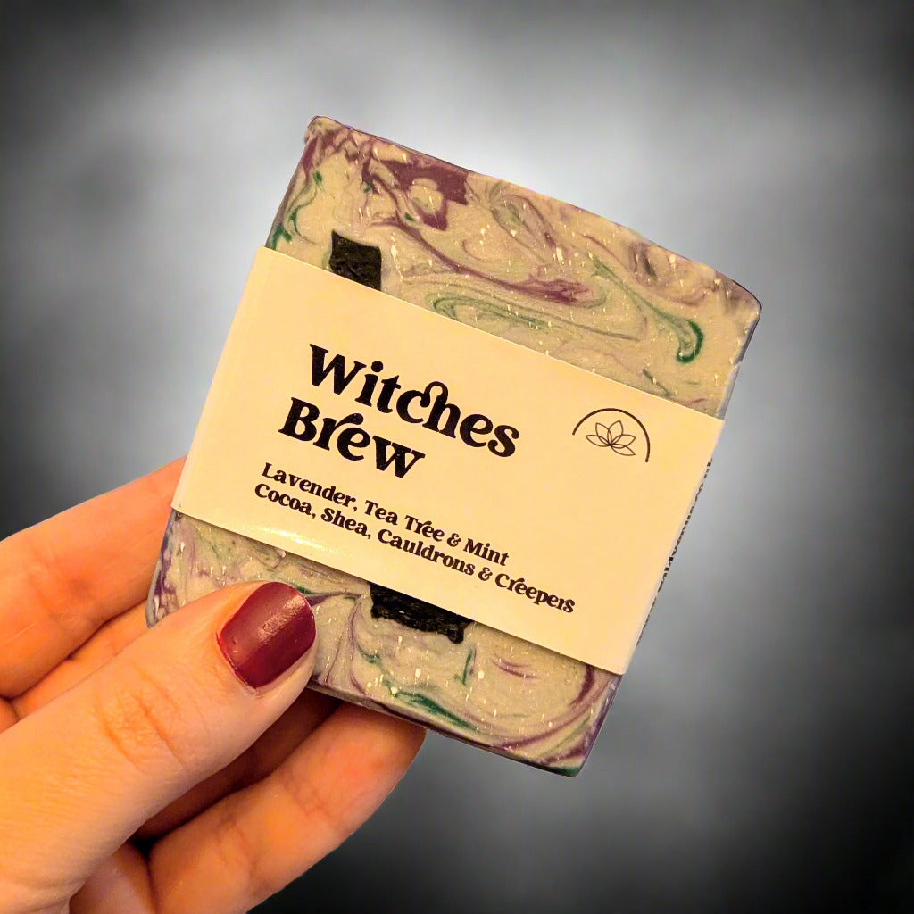 Holding a bar of Witches Brew soap with a label on against a grey and white background.