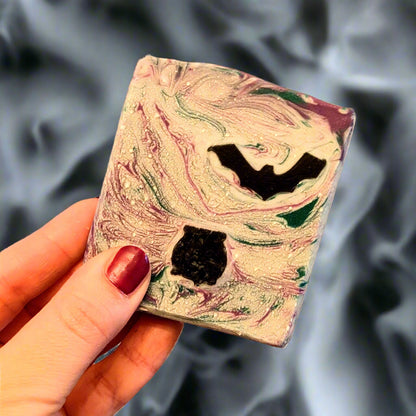 Holding a bar of unwrapped soap which has a bat and a cauldron in. The soap is held against a spooky and smoky black background.