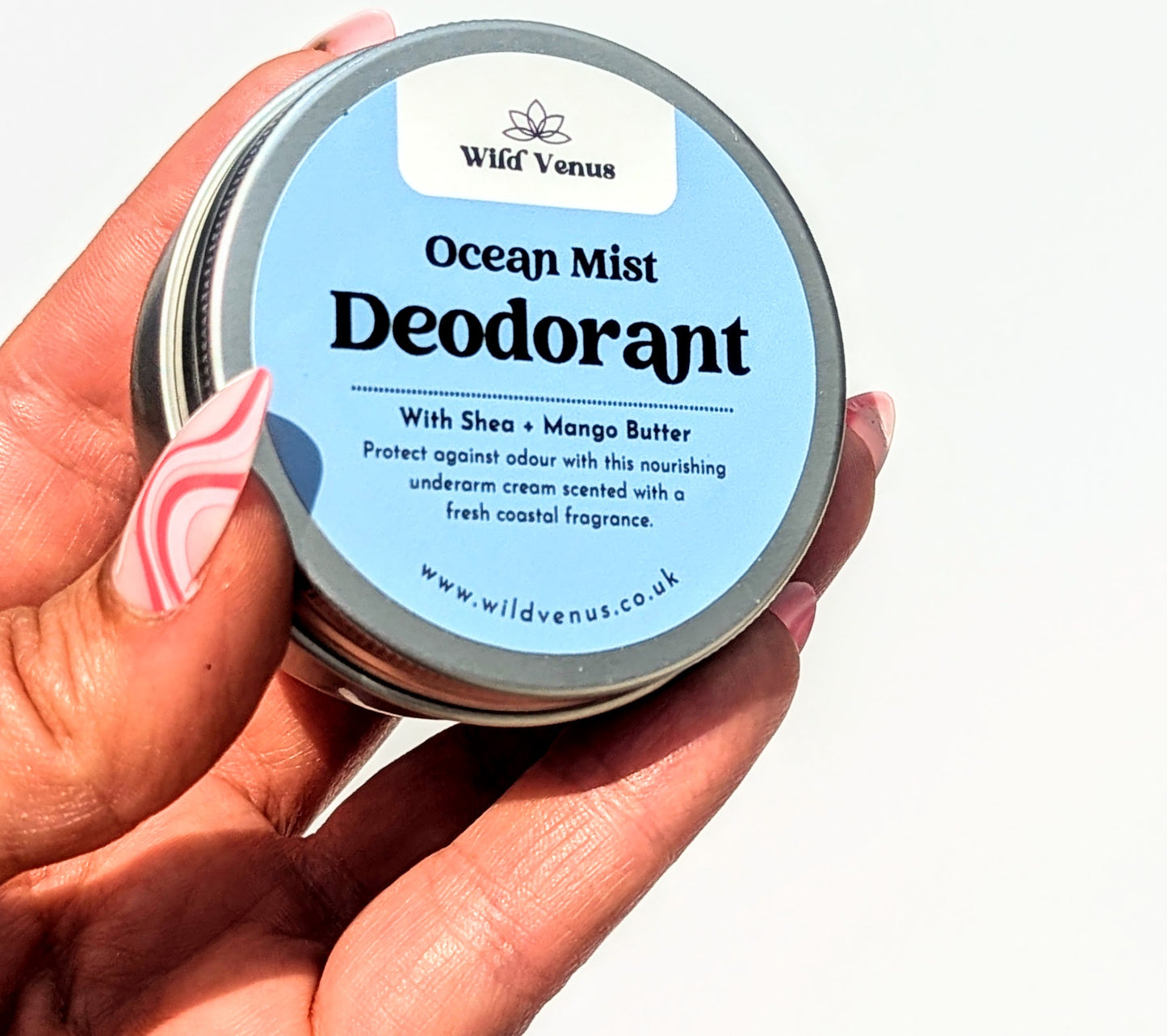 A manicured hand holds up a tin of Ocean Mist scented deodorant cream against a white background. 