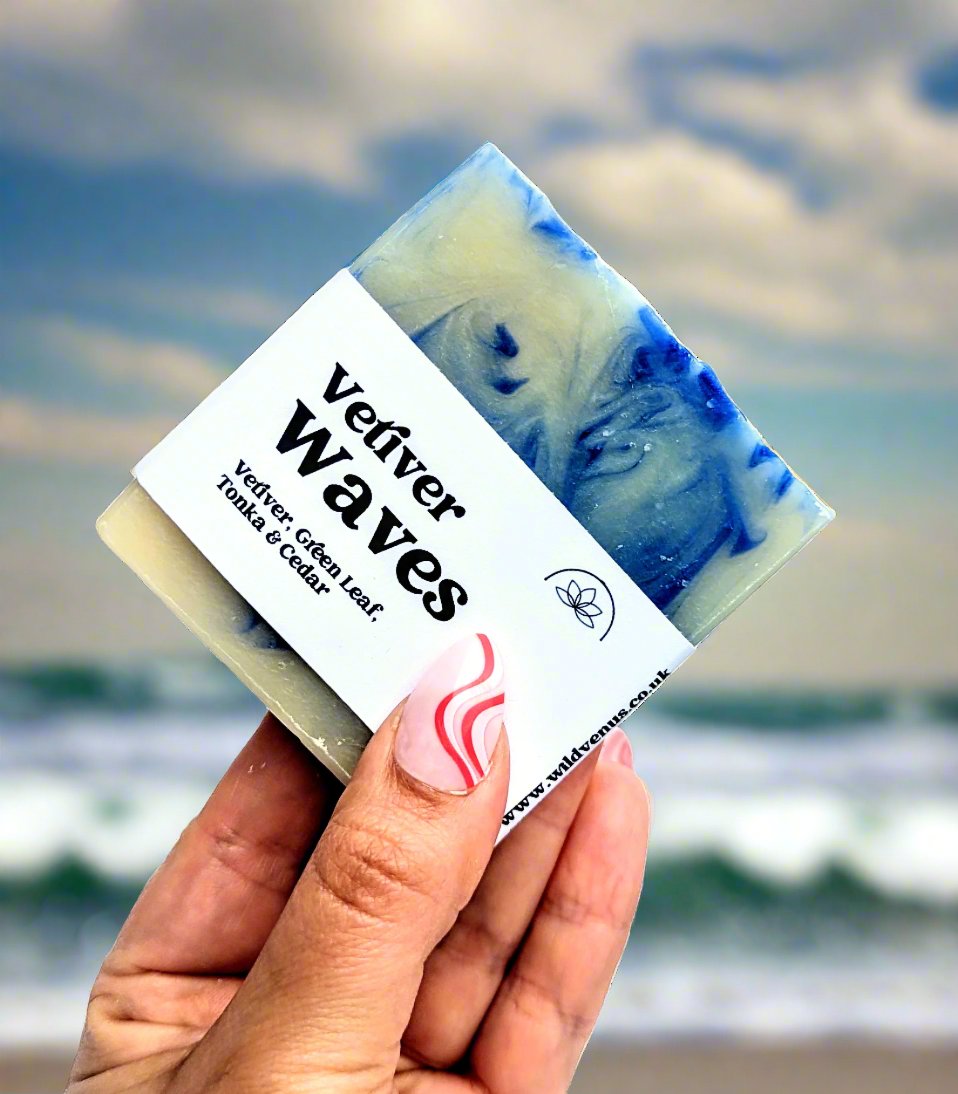 Holding a bar of Vetiver Waves handmade soap at the beach with waves and a blue sky in the background.